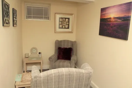 A grey wing-back armchair beside a window in the Uxbridge consulting room, with a soft sunset canvas on the wall.